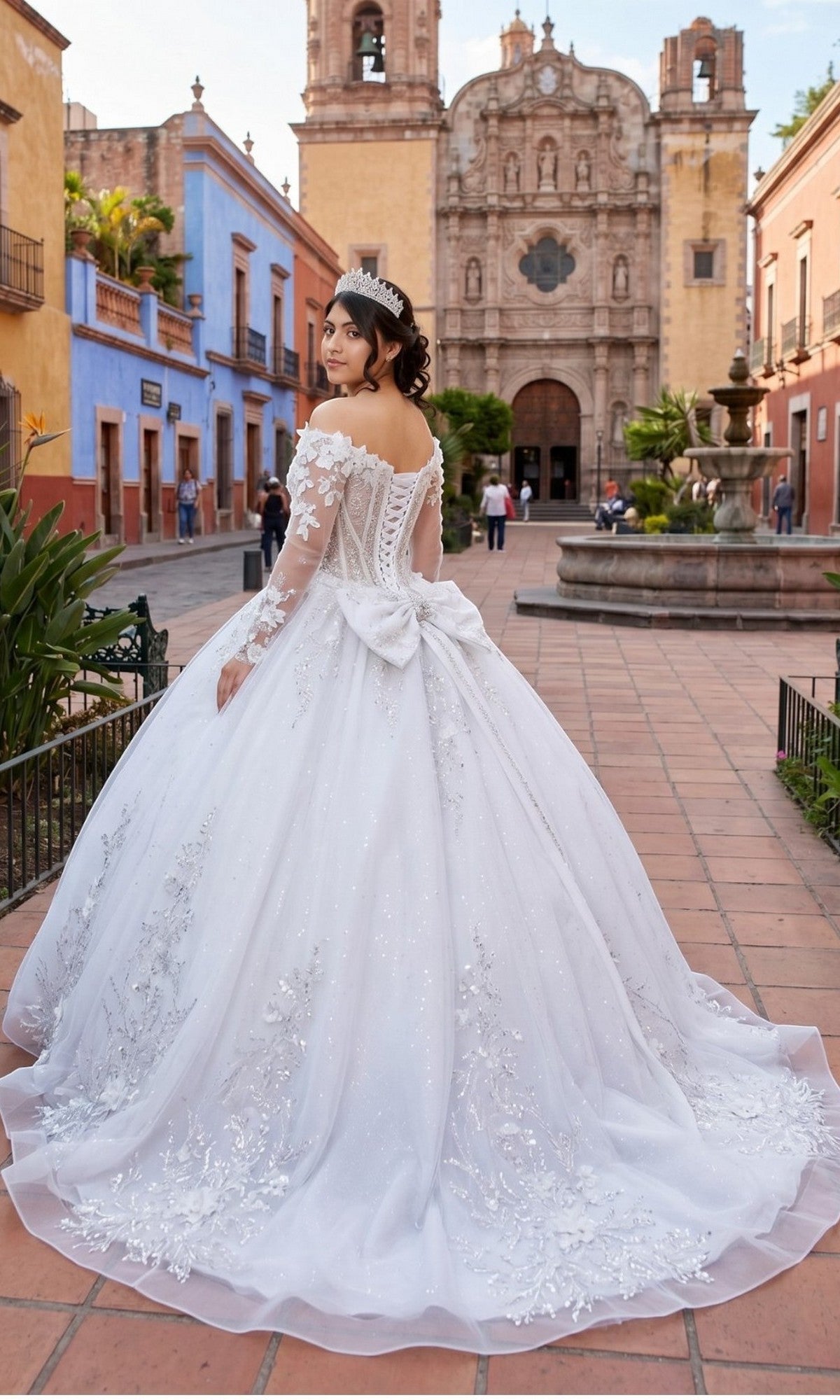 A young woman wearing the Cinderella Couture 8325J long formal dress—a white glitter tulle ball gown with a delicate corset bodice and a tiara—stands on a cobblestone street, framed by colorful buildings and a historic church in the background.
